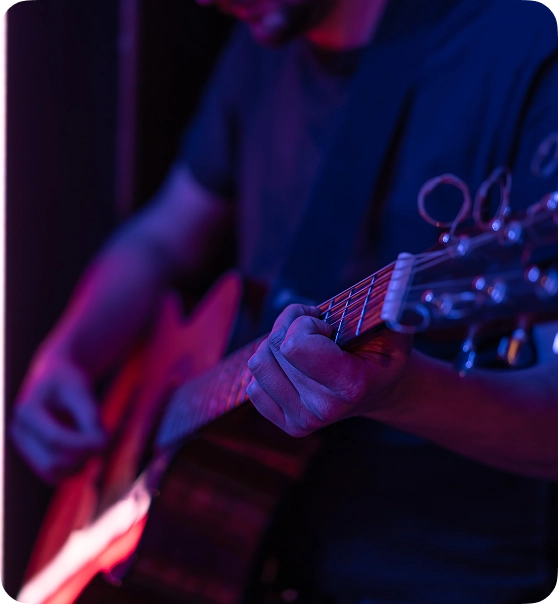 Een persoon bespeelt een akoestische gitaar onder fel paars en roze licht, wat een stemmige, artistieke sfeer creëert. De aandacht is gericht op de gitaar en de handen.
