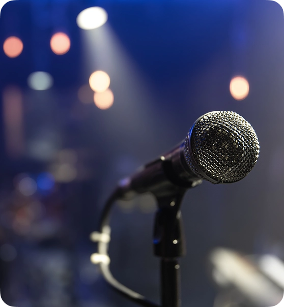 Close-up van een microfoon op een statief in een schemerig verlichte podiumomgeving. Felle lichten en wazige vormen op de achtergrond creëren een energieke sfeer.